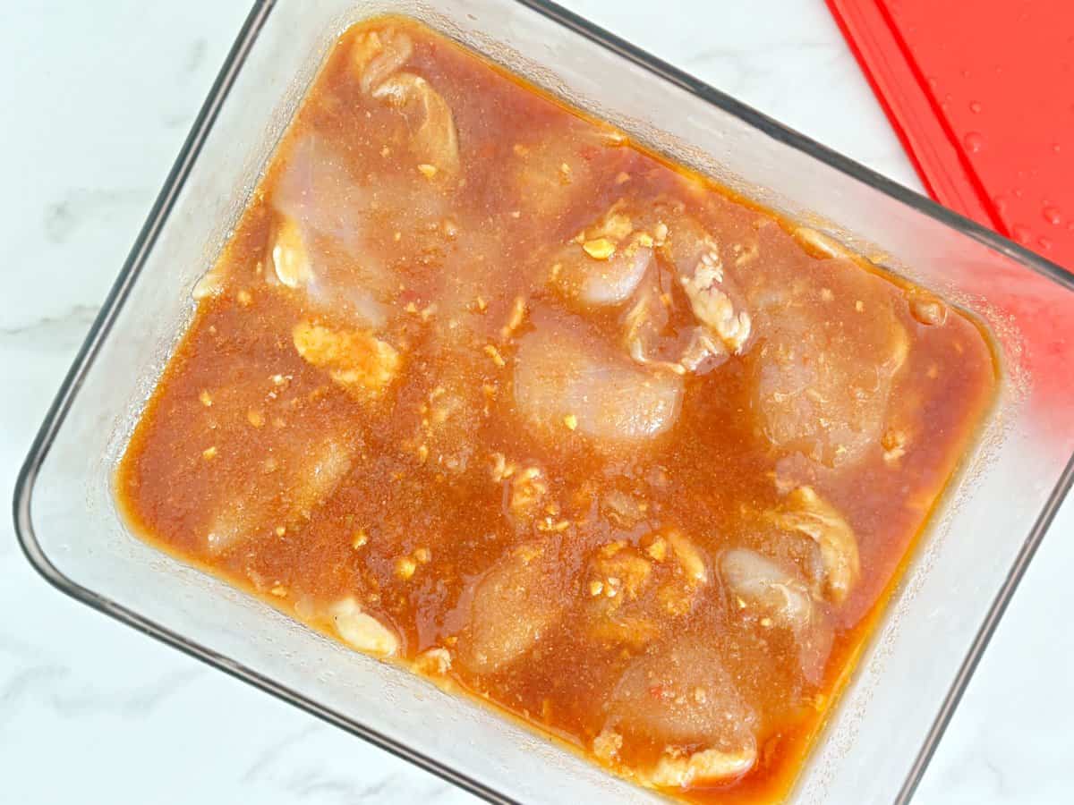 Down view of chicken thighs marinating in clear glass container.