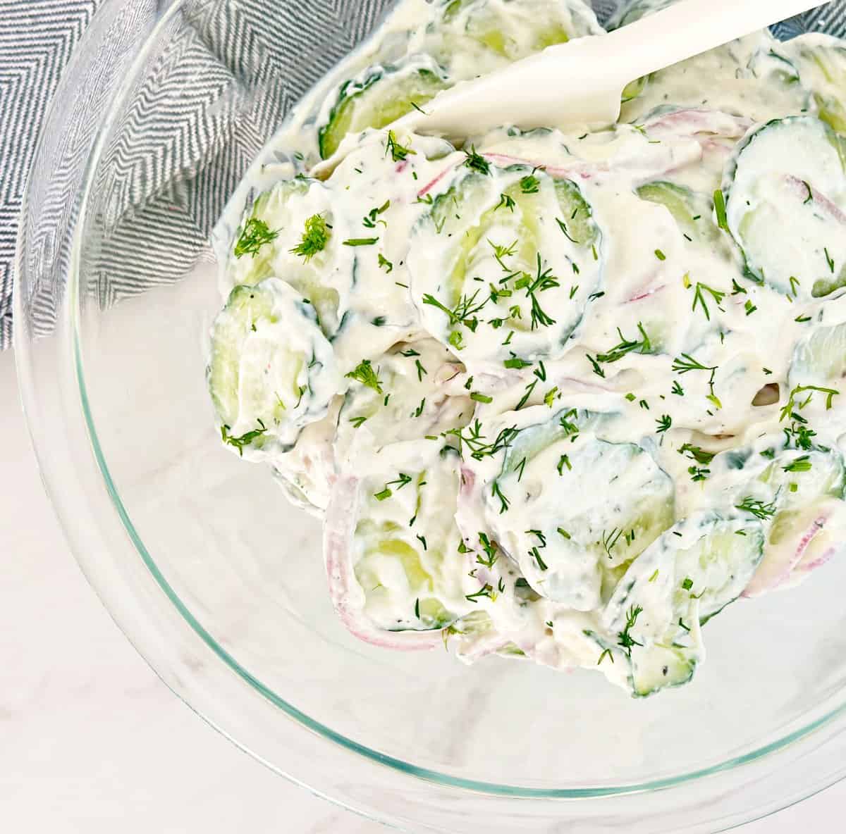 Down view of clear mixing bowl with creamy cucumber salad and white spatula.