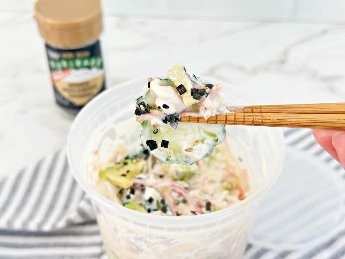 Chopstick holding a portion of sushi cucumber salad from a deli container.