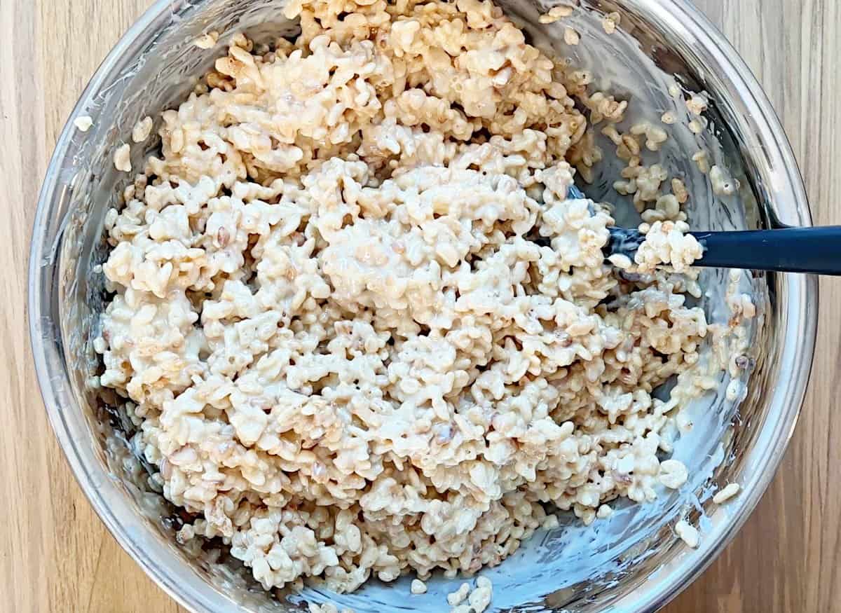 A silver bowl with rice krispies mixture and a black rubber spatula in it.