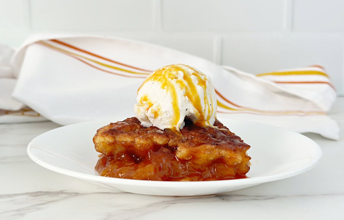 Peach cobbler topped with vanilla ice cream and caramel sauce on white plate.