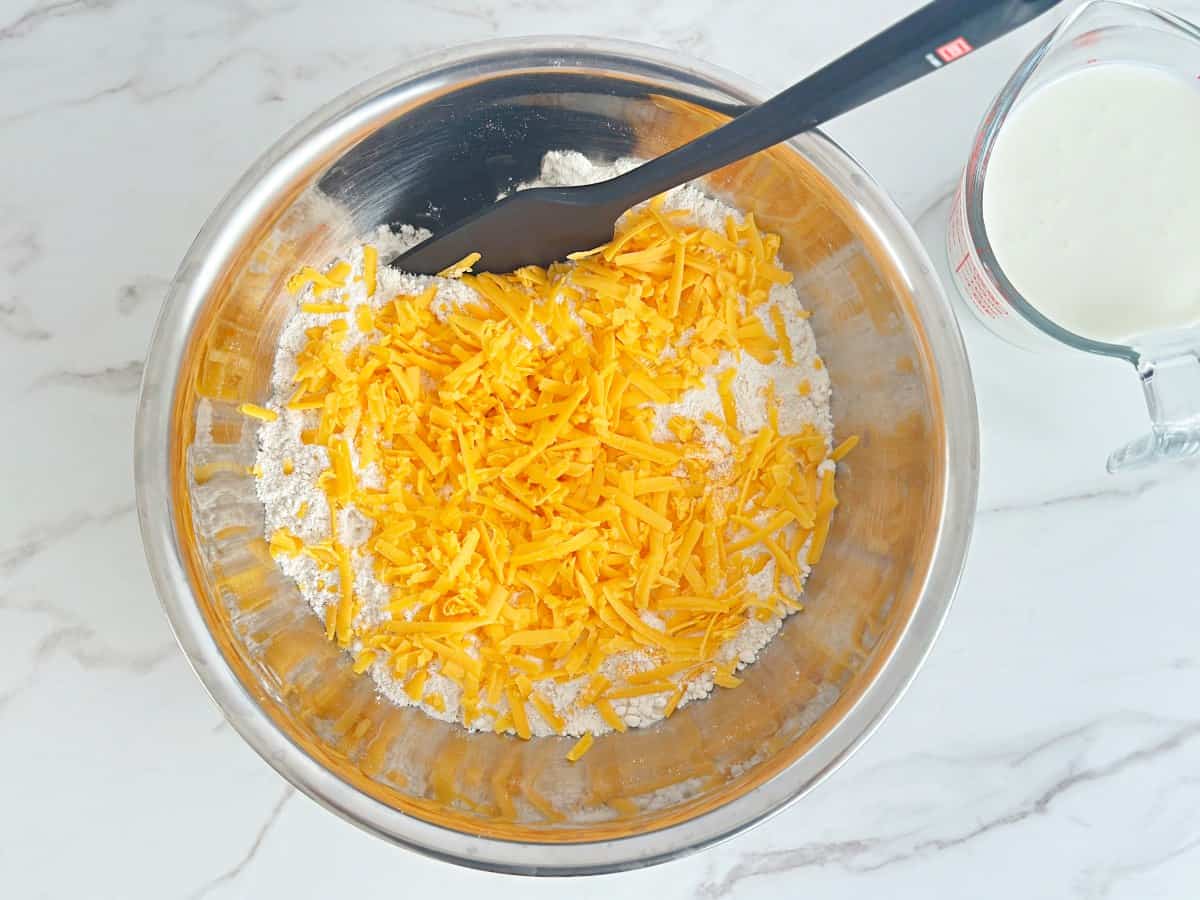 Silver mixing bowl with dry mixture and shredded cheese, next to a measuring cup with buttermilk.