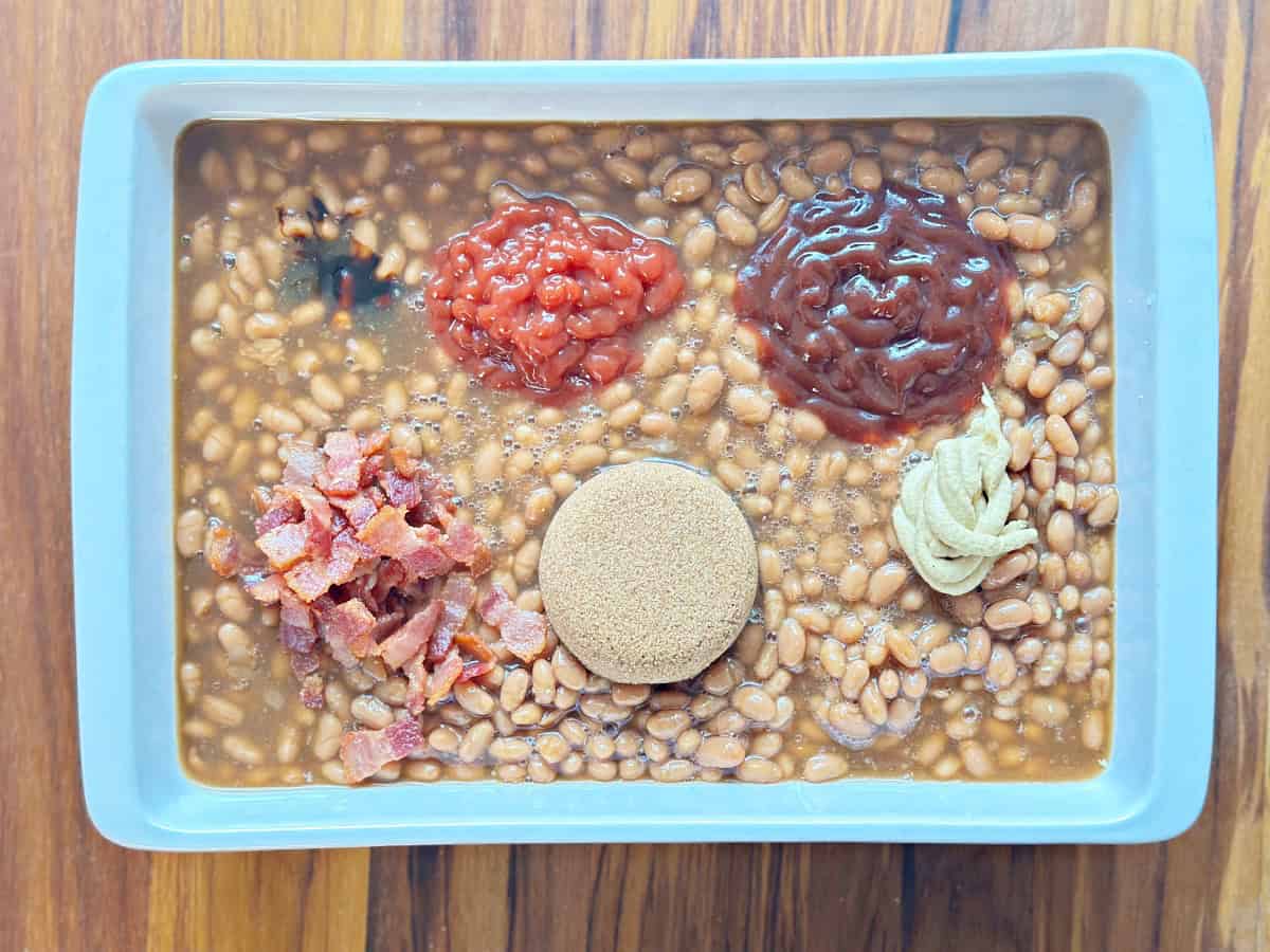 Down view of white baking dish with bean mixture topped with brown sugar, bacon, bbq sauce, ketchup, mustard, and molasses.