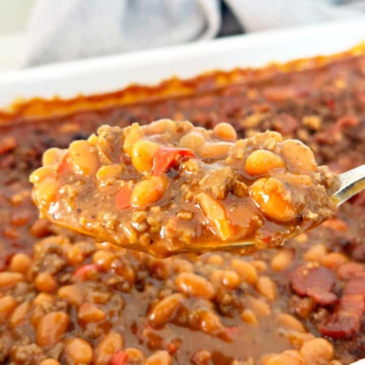 A spoonful of cowboy baked beans is being held up on a spoon under is a casserole dish.