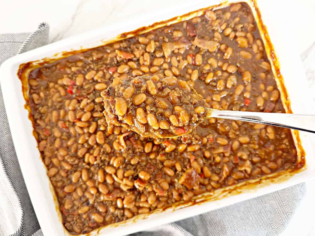 Cowboy baked beans with ground beef on silver spoon. Under is a white baking dish with more baked beans.