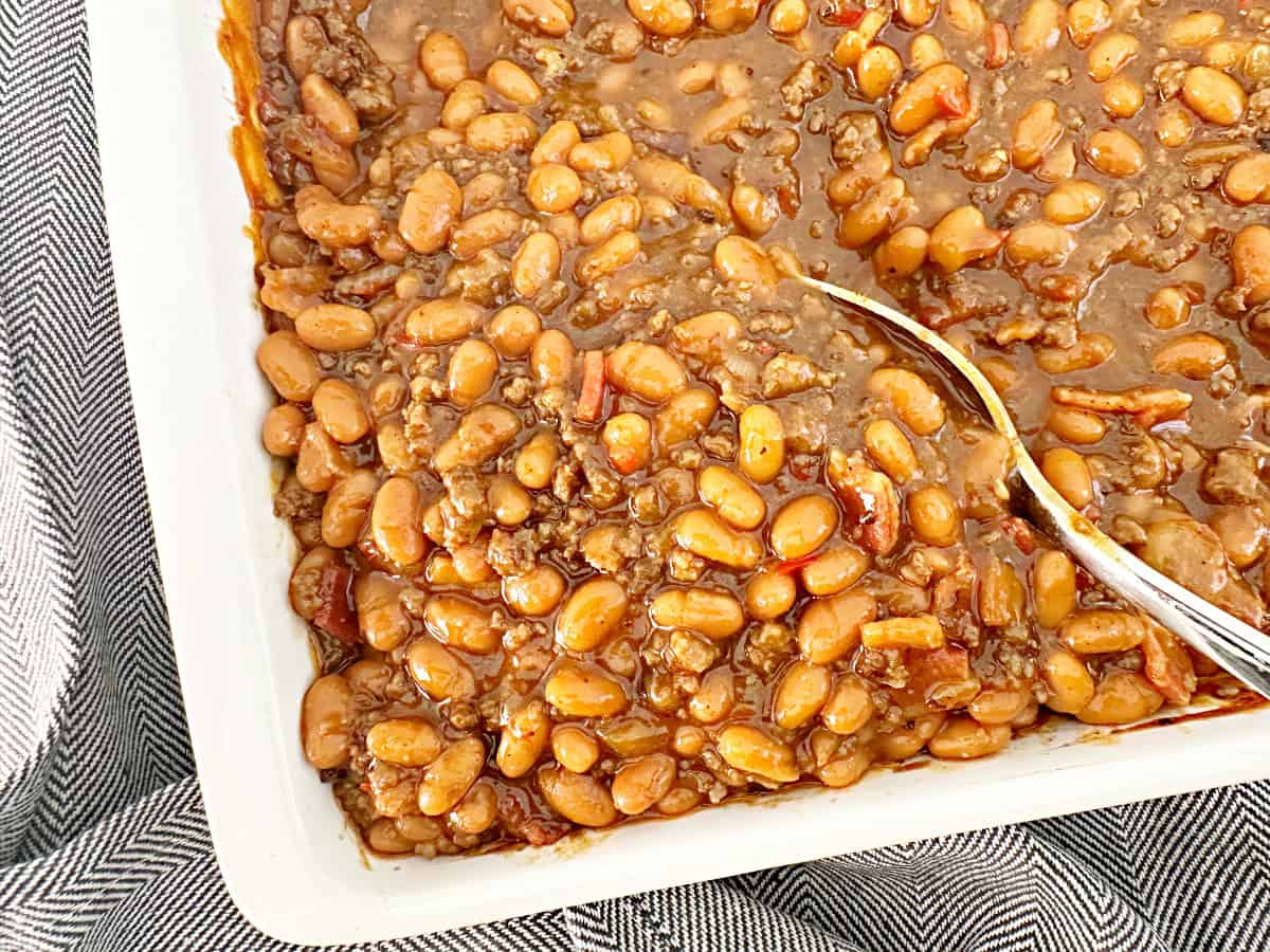 Down view of easy cowboy baked beans in white baking dish with silver serving spoon.