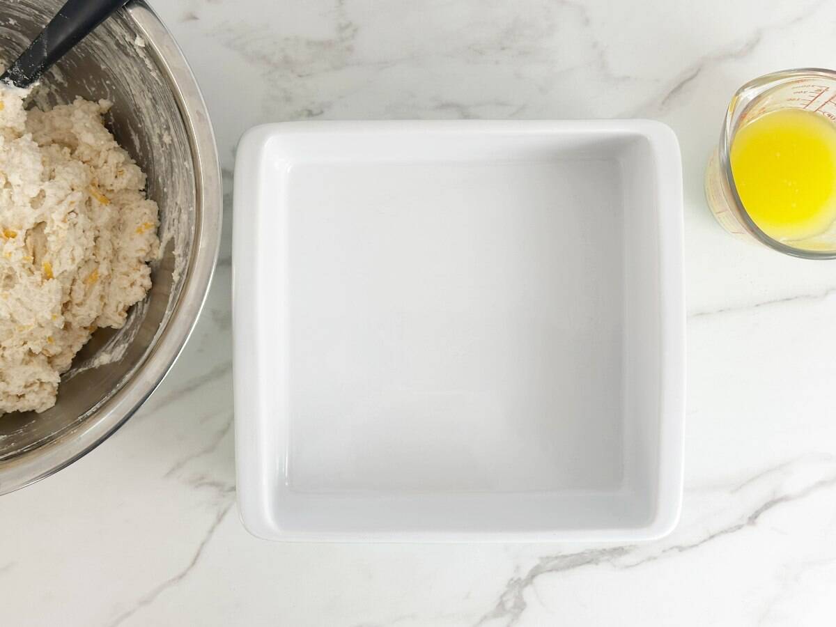 A white baking dish next to a mixing bowl with biscuit mixture and a measuring cup with melted butter.