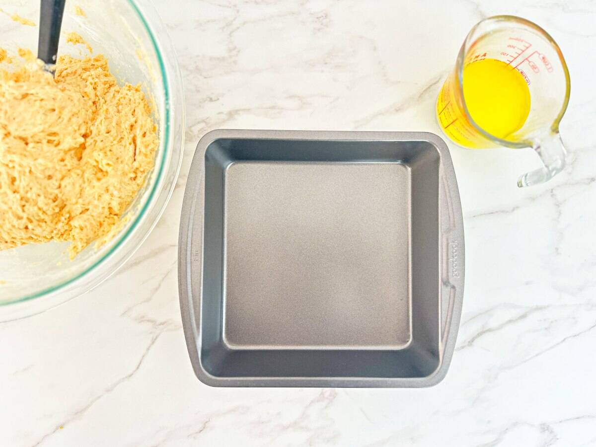 A metal baking dish next to a mixing bowl with biscuit mixture and a measuring cup with melted butter.