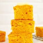 Three slices of Jiffy sweet potato cornbread stacked on each other, topped with a cube of butter. Surrounded by more cornbread on wire rack.