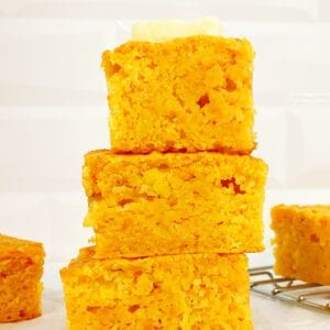 Three slices of Jiffy sweet potato cornbread stacked on each other, topped with a cube of butter. Surrounded by more cornbread on wire rack.
