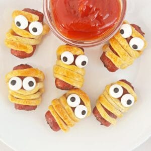 Mini mummy dogs on a white plate next to a small clear bowl with ketchup.