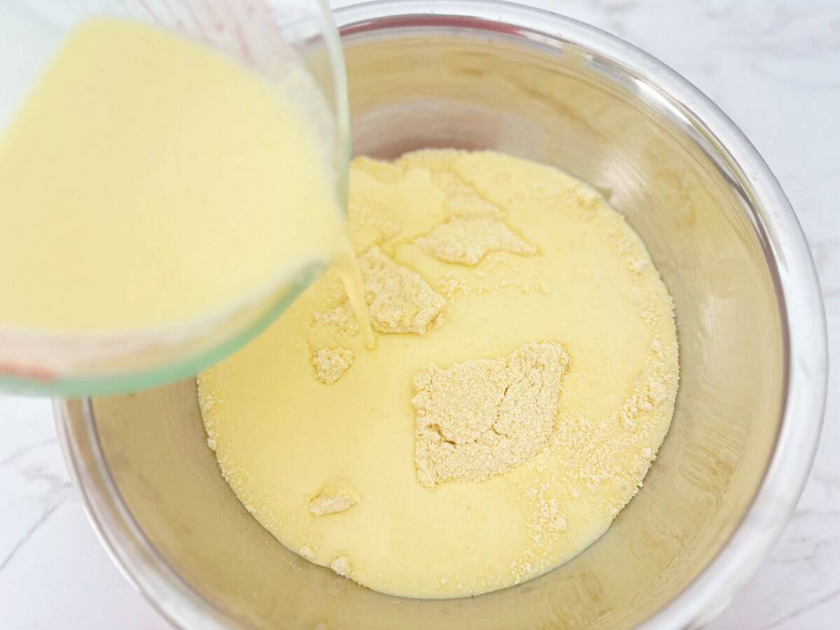 Wet ingredients being poured into Jiffy muffin mix in a silver mixing bowl, down view.