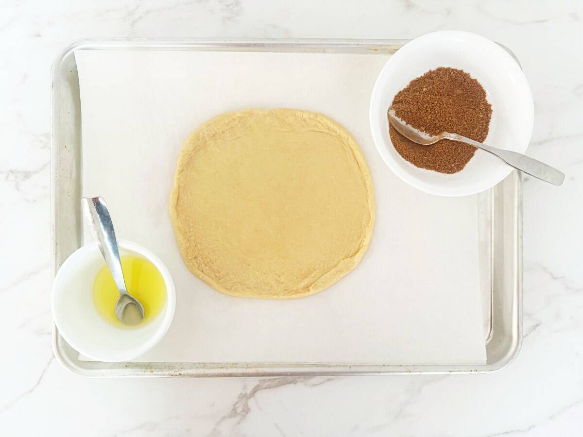 Crescent roll pizza crust on parchment-lined sheet pan. Next to two mixing bowls with cinnamon sugar and melted butter, down view.